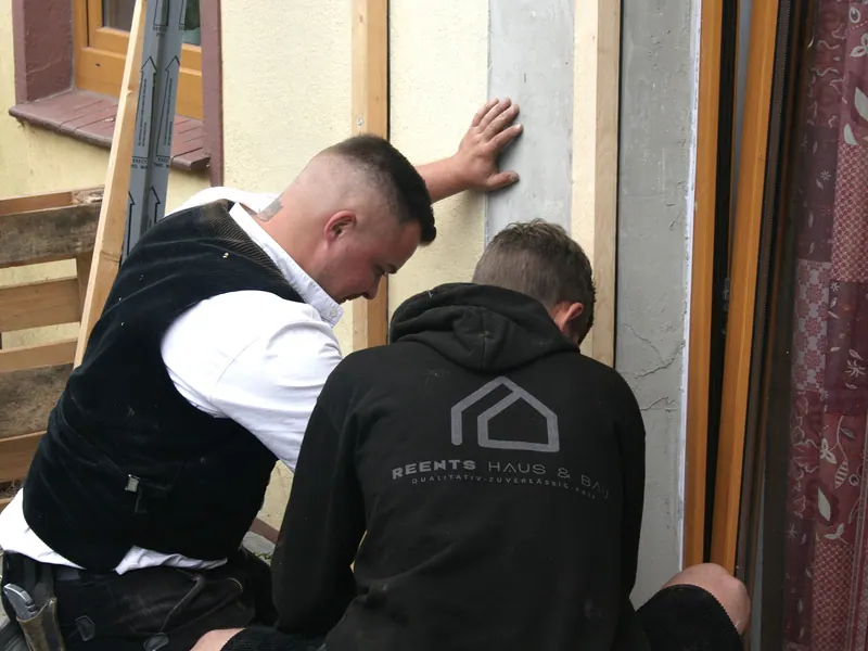 Zwei Handwerker setzen einen Holzrahmen für eine Tür in die Fassadenöffnung