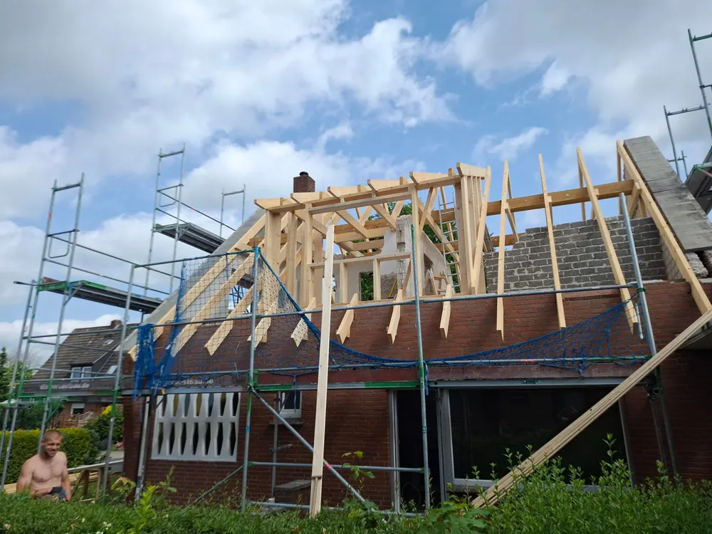 Frisch aufgerichteter Dachstuhl aus hellem Holz auf einem Klinkerhaus, rundum Gerüst mit grünem Schutznetz